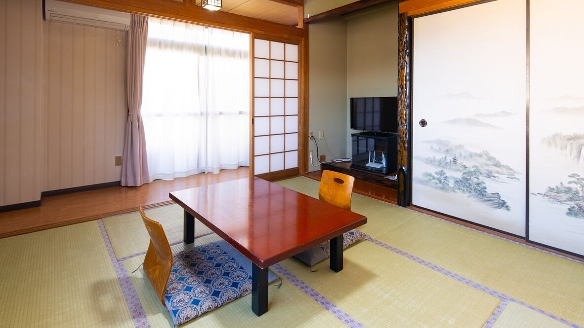 A traditional Japanese room with tatami flooring, a low wooden table, a chair, a television, and a sliding door with a landscape painting.