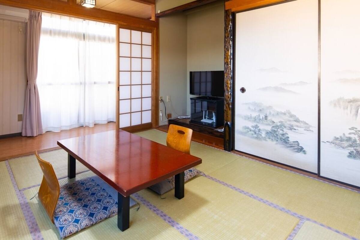 A traditional Japanese room with tatami flooring, a low wooden table, a chair, a television, and a sliding door with a landscape painting.