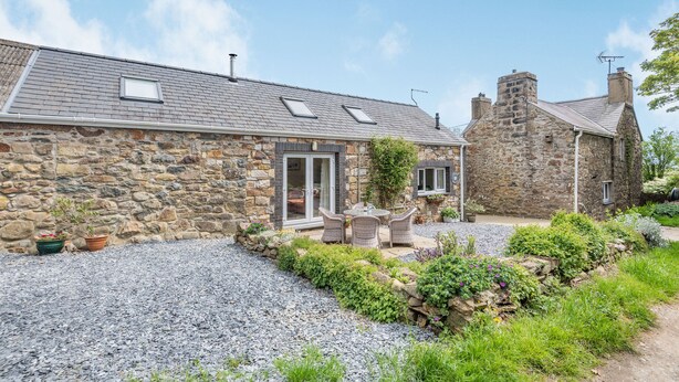 Une maison en pierre avec une allée en gravier et un patio aménagé avec des meubles en osier.