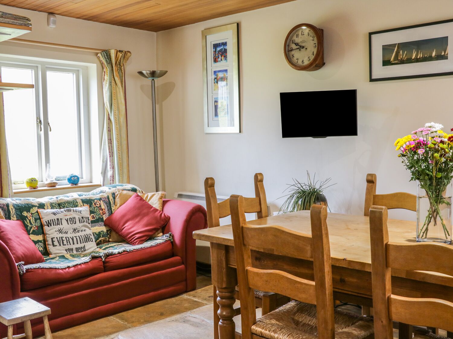 A cozy living room with a red sofa, wooden dining table, and chairs. A flat-screen TV is mounted on the wall above the table.