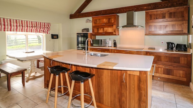 A kitchen with wooden cabinets, a large island with stools, a window with a view, and a dining table with chairs. - Rosies Cottage (Bushmills)