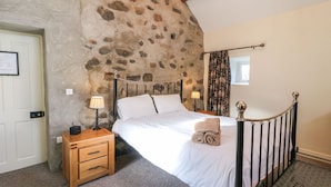 A bedroom with a stone wall, a bed with a metal frame, a wooden nightstand, a lamp, and a window with curtains. - Rosies Cottage (Bushmills)
