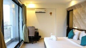 A hotel room with a bed, a window with curtains, a wall-mounted air conditioner, and a small table with a towel. - Pallavi Ávida (Panvel)
