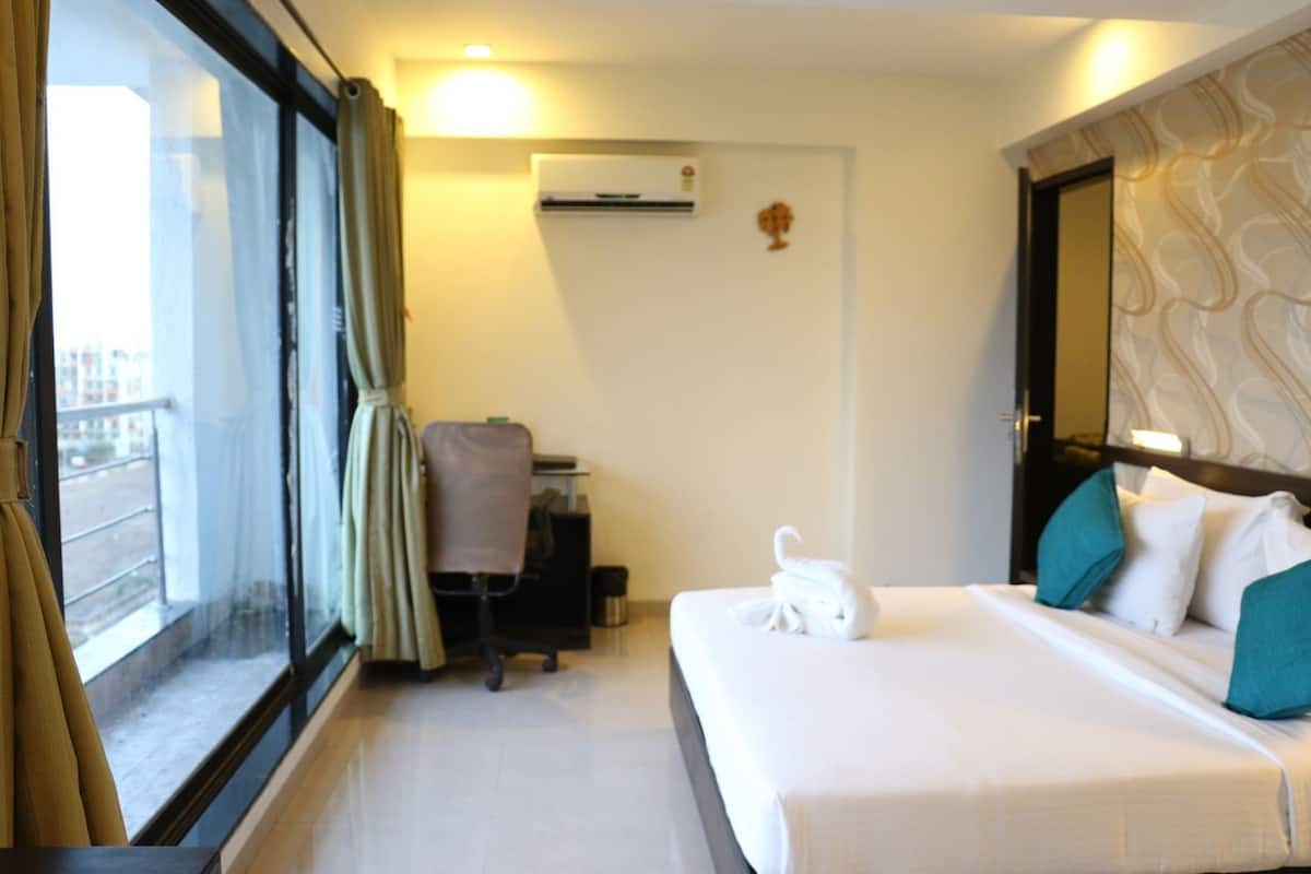 A hotel room with a bed, a desk, a chair, a window with curtains, an air conditioning unit, and a wall-mounted decoration.