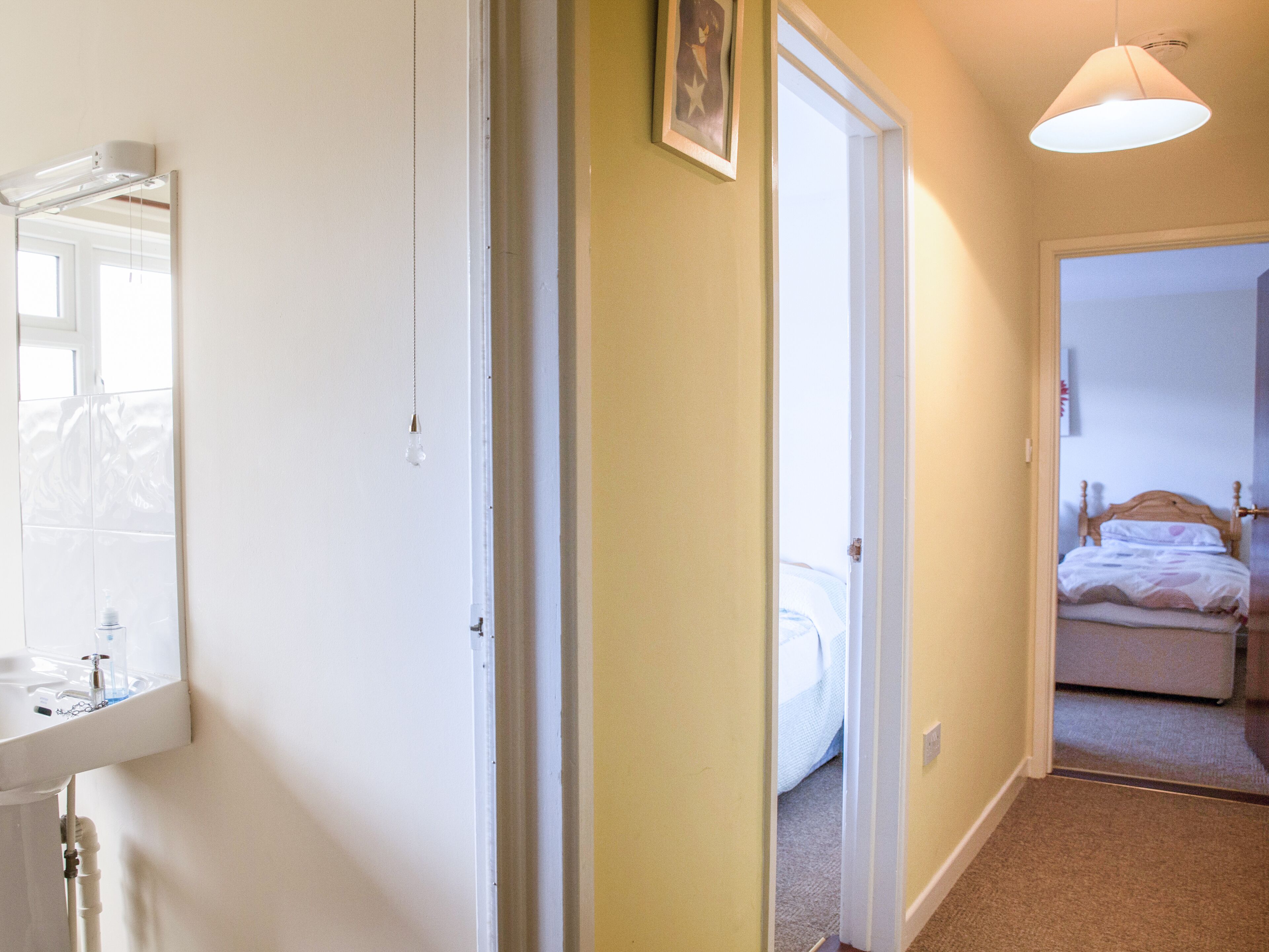 A hallway leading to a bedroom with a bed, a bedside table, and a nightstand. There is a bathroom with a sink and mirror visible to the left.