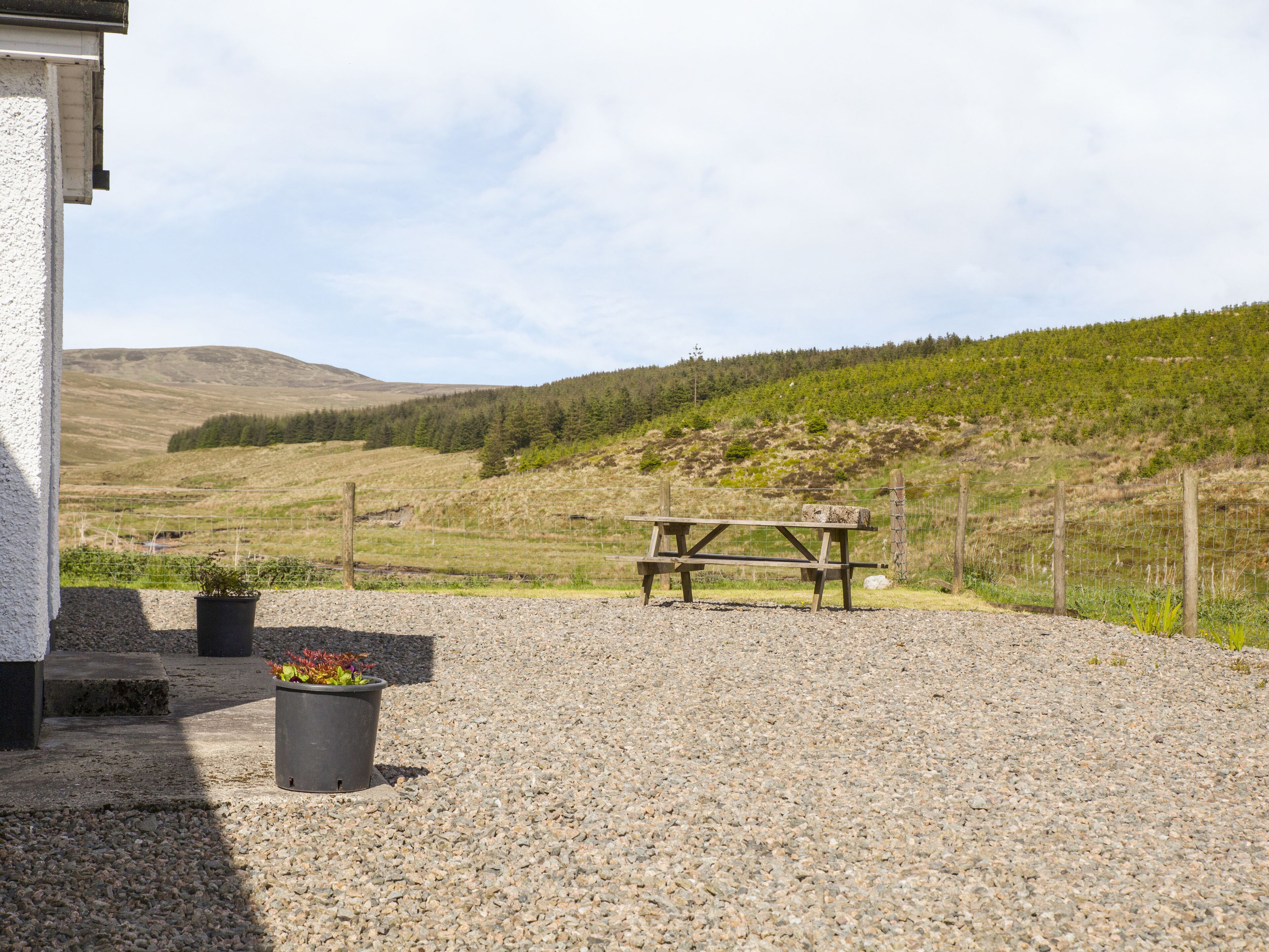 A gravel patio with a wooden table and chairs, surrounded by a fence and a view of a hilly landscape.