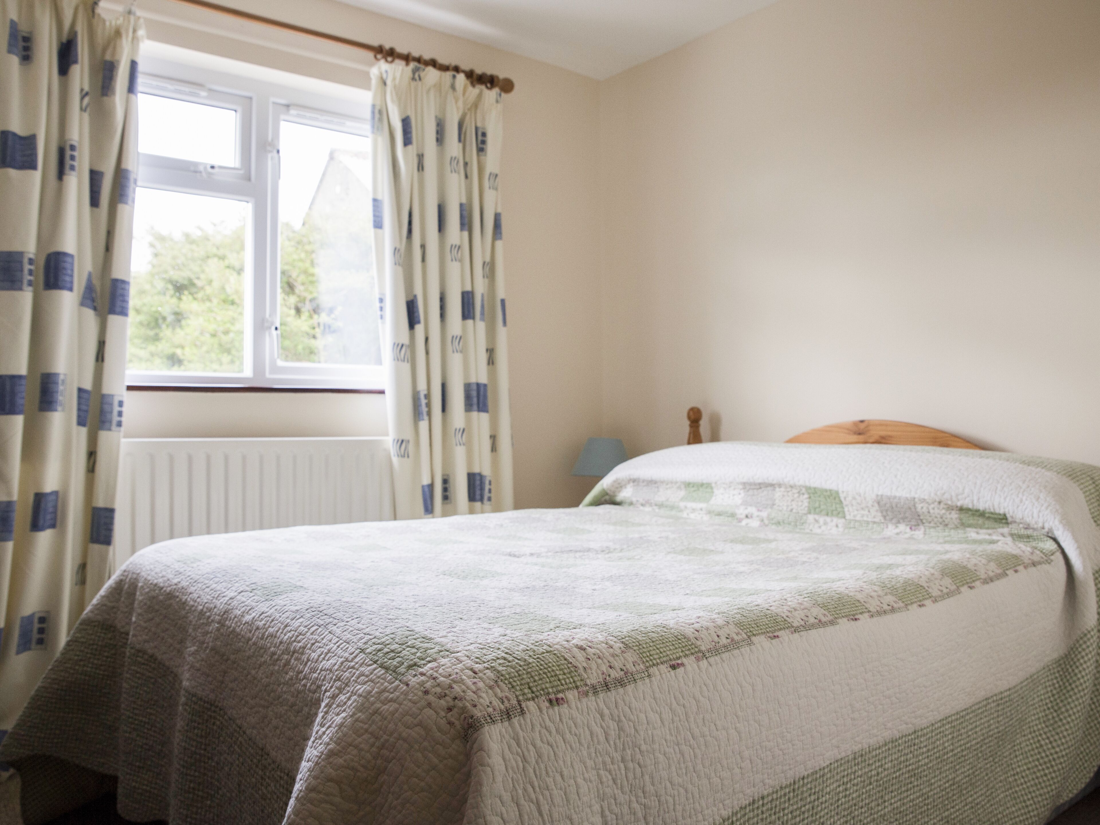 A bedroom with a bed, a window with curtains, a radiator, and a nightstand with a lamp.