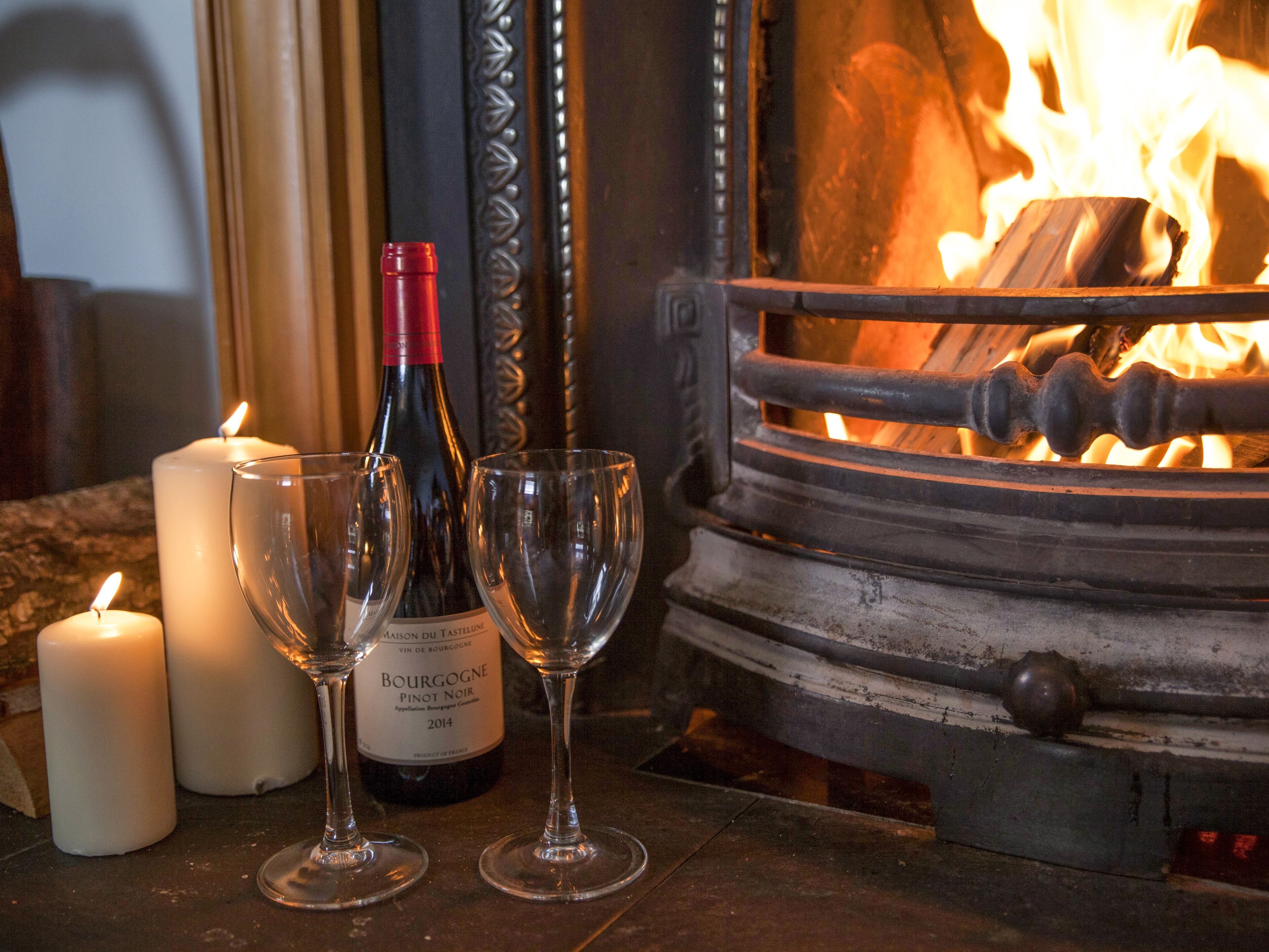 Una chimenea con leña ardiendo, dos velas y una botella de vino con dos copas.