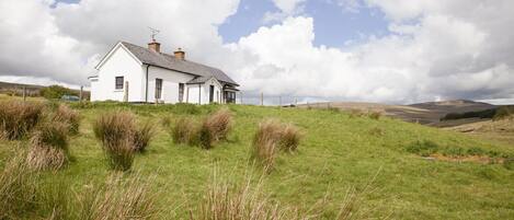 A white house with a sloped roof and chimneys, surrounded by grassy fields and hills in the background.