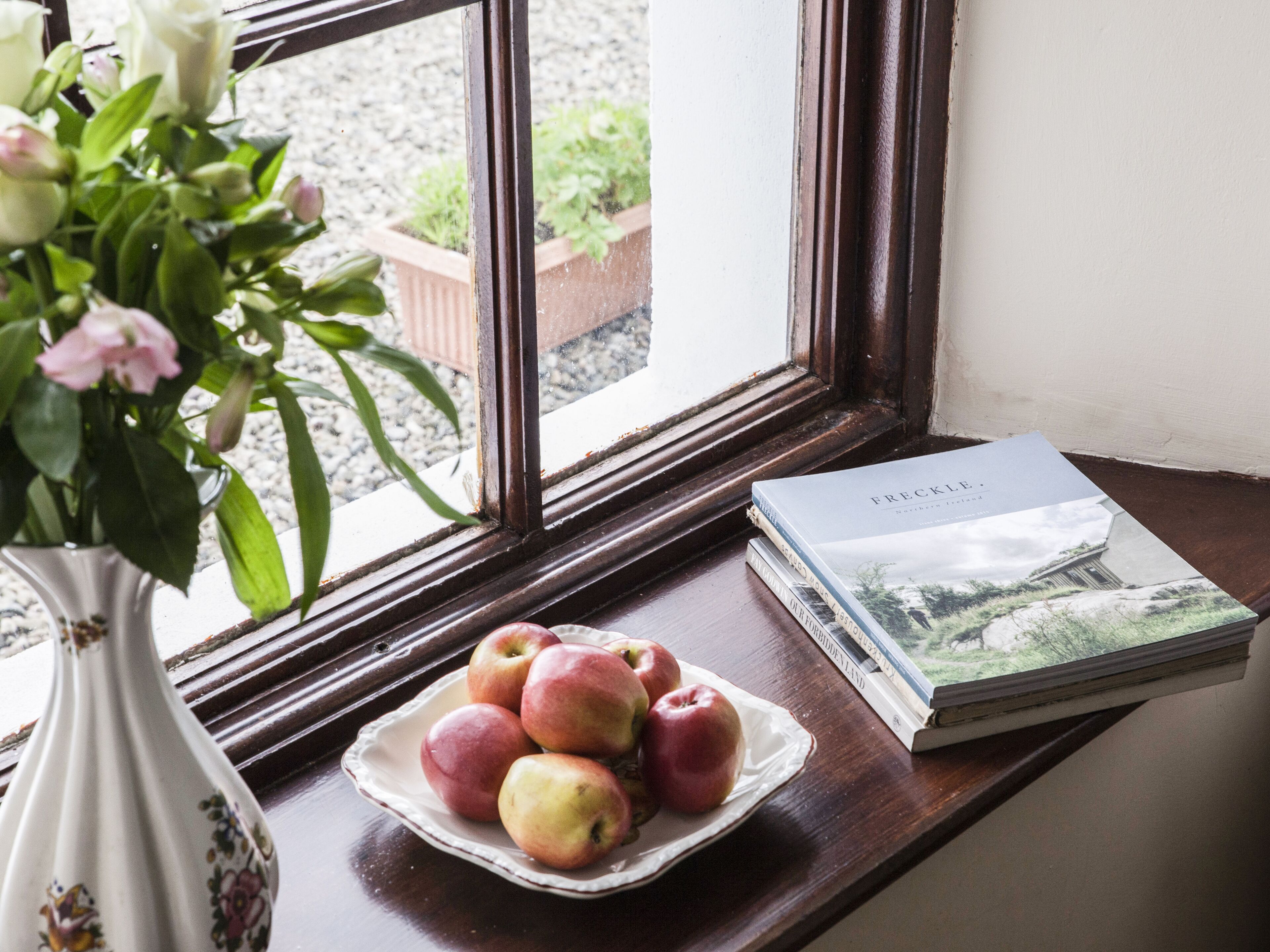 Una ventana con vistas al verdor exterior, un jarrón con flores, un plato de manzanas y una pila de libros en el alféizar.