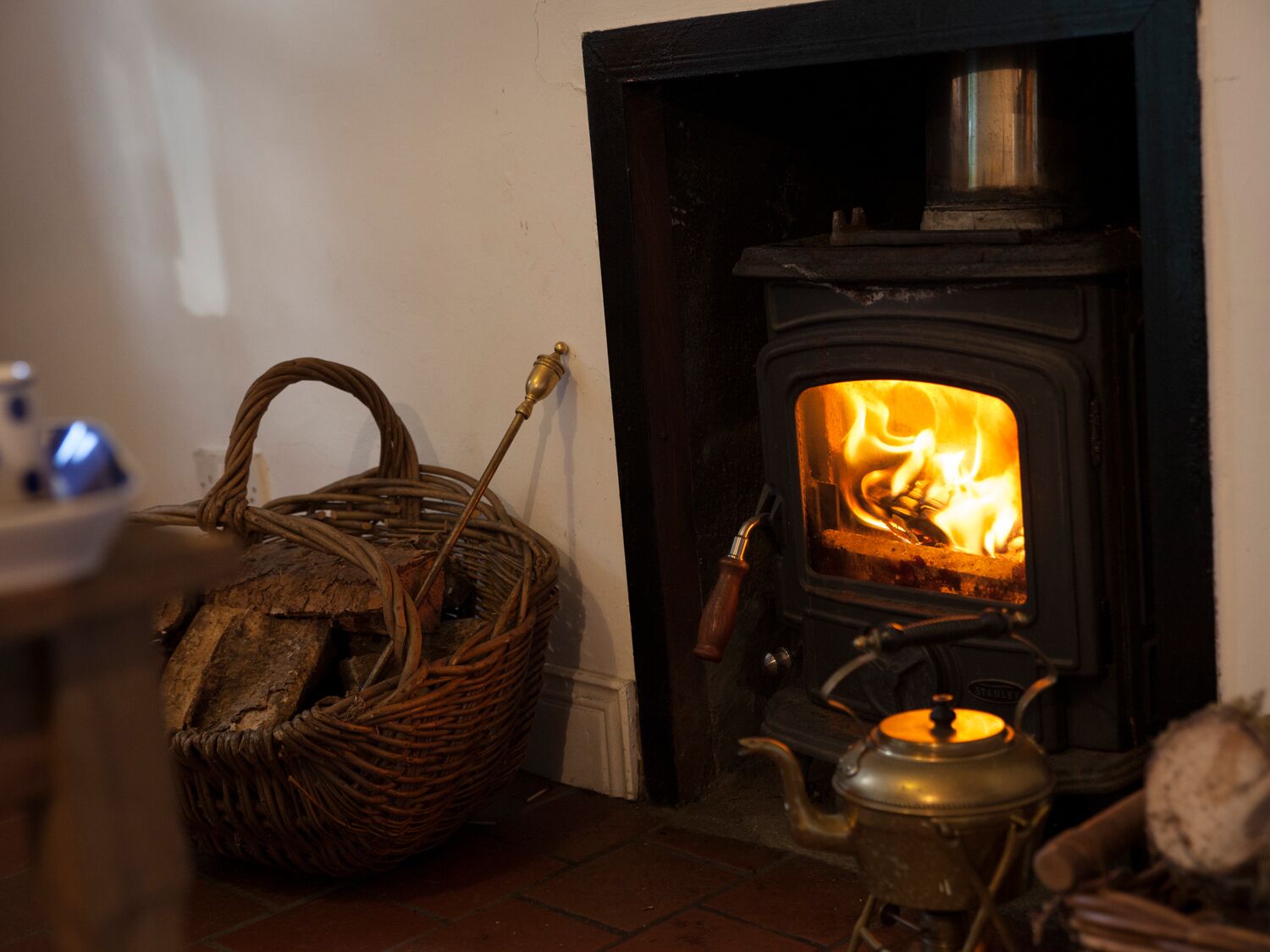 A cozy room with a wood-burning stove, a wicker basket, and a brass kettle.