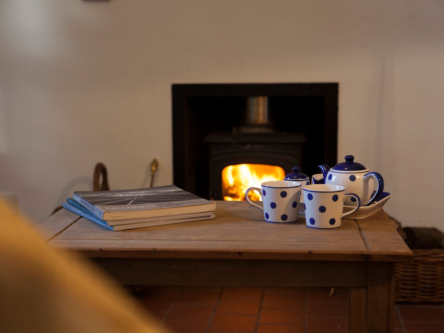 A wooden table with a stack of books, a teapot, and two mugs in front of a fireplace.