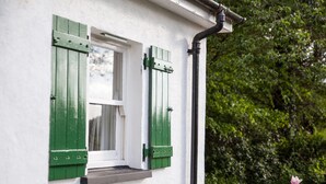 A white building with a window featuring green shutters, a small step leading to the entrance, and a black pipe running along the wall. - Mr Mcgregors' Cottage (Omagh)