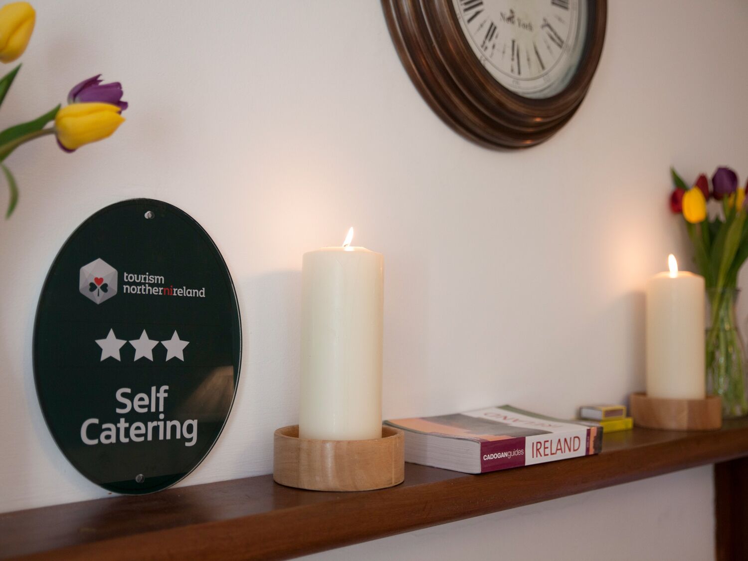 A self-catering sign, two lit candles, a clock, and a book titled 'Ireland' on a shelf.