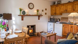 A cozy living room with a fireplace, wooden cabinets, a dining table set for four, and a kitchen area with modern appliances. - Mr Mcgregors' Cottage (Omagh)