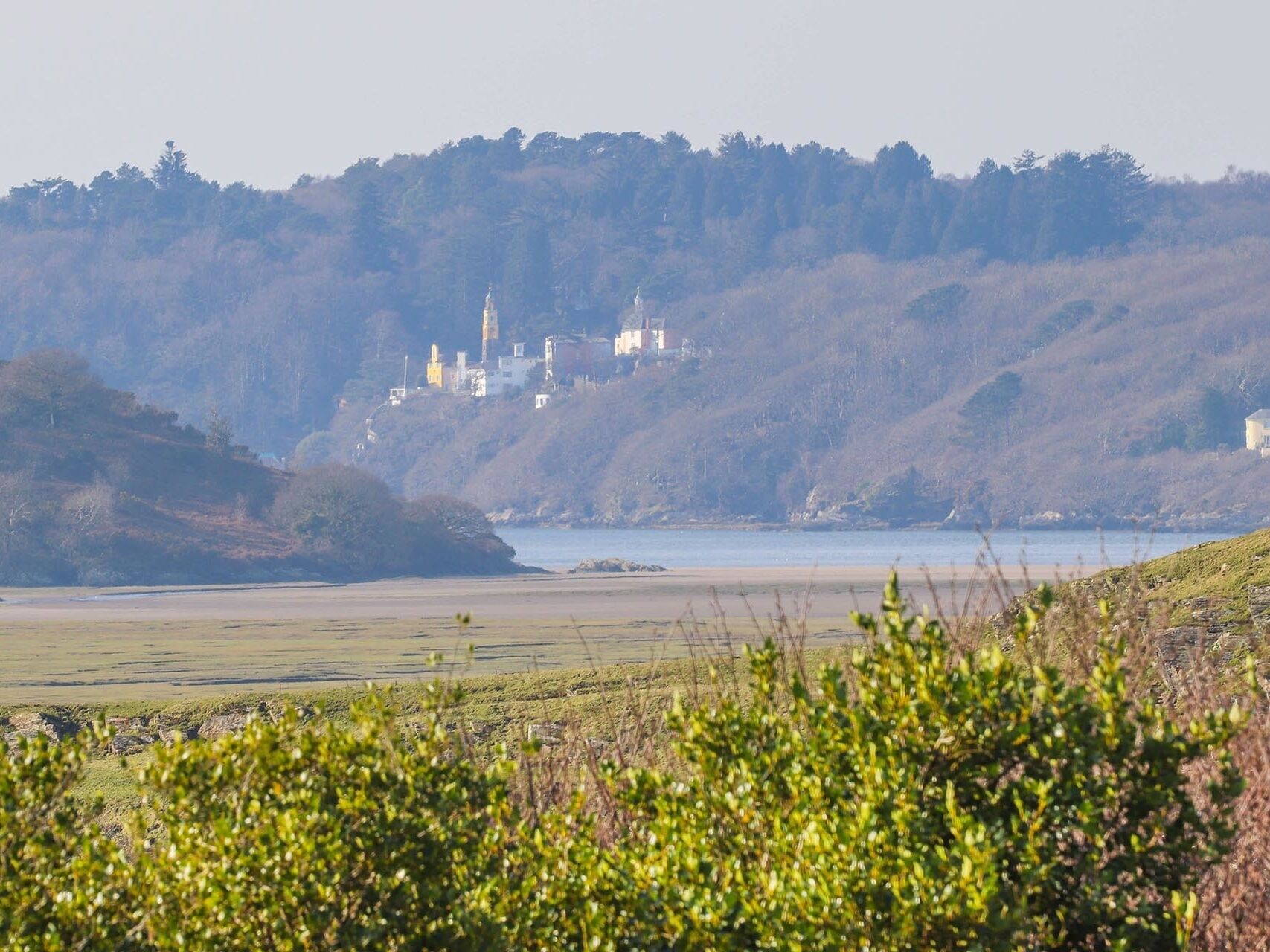A coastal landscape with a historic building complex on a hill, surrounded by greenery and a calm sea.
