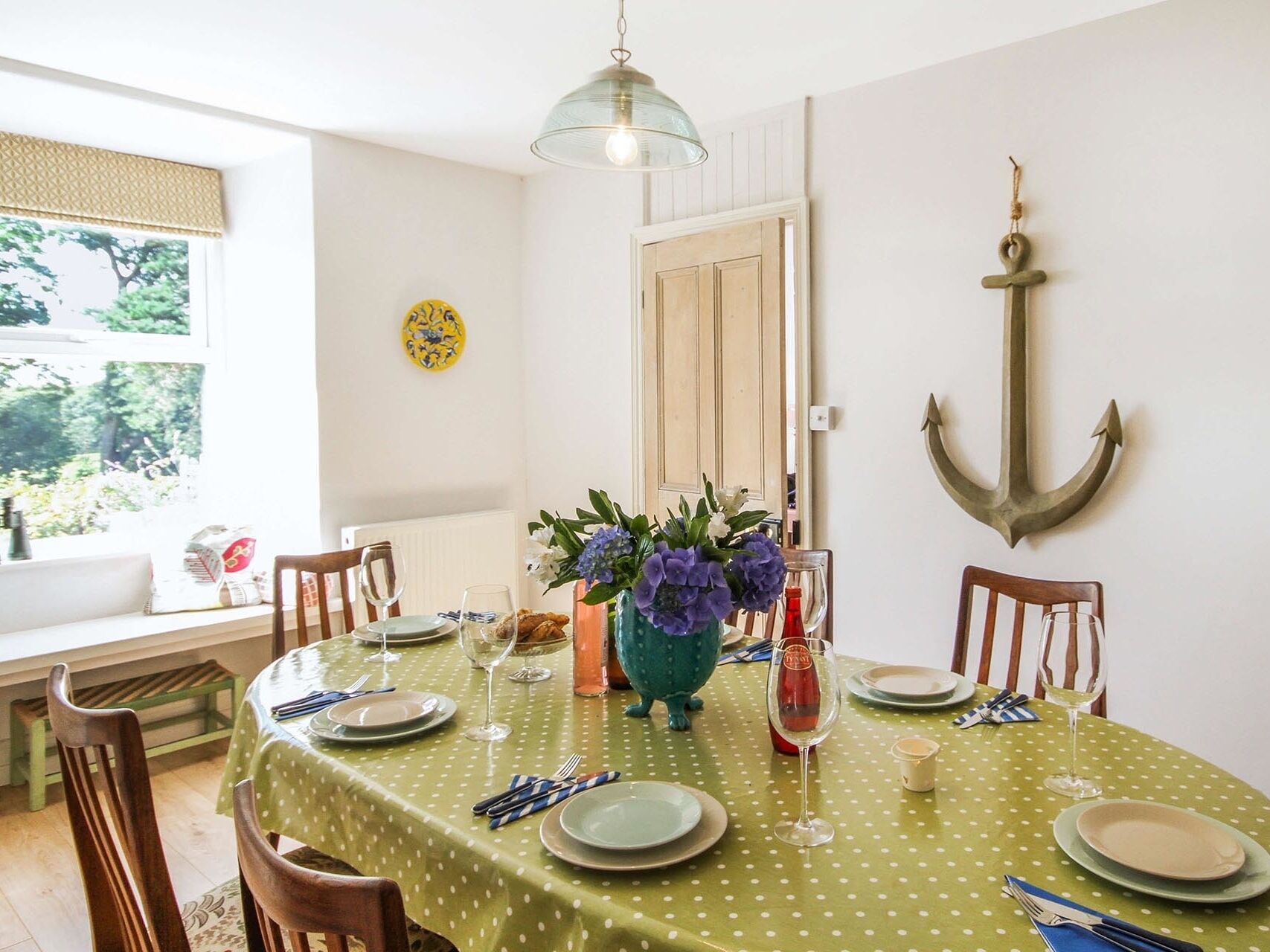 A dining area with a round table set for four, covered with a green tablecloth. There are chairs around the table, a vase with flowers, and a wall-mounted anchor decoration.