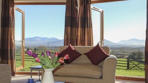 Une pièce chaleureuse avec un canapé placé face à une grande fenêtre, une table basse ornée d’un vase de fleurs, et une vue sur un paysage verdoyant avec des montagnes.