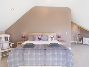 A bedroom with a sloped ceiling, a bed with a plaid bedspread, two nightstands with lamps, a chair, and a small table with a vase. - Dol Fraint (Gaerwen)