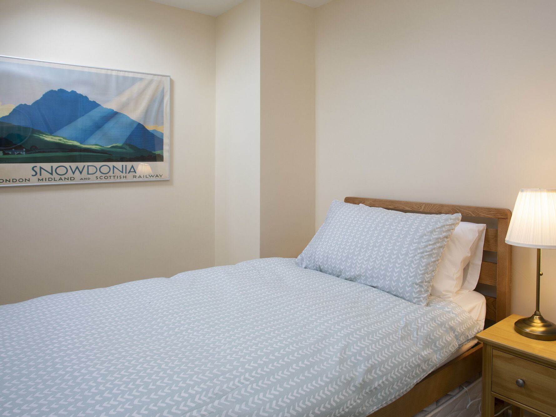 A neatly made bed with a patterned bedspread, a wooden headboard, a bedside lamp, and a poster of Snowdonia on the wall.