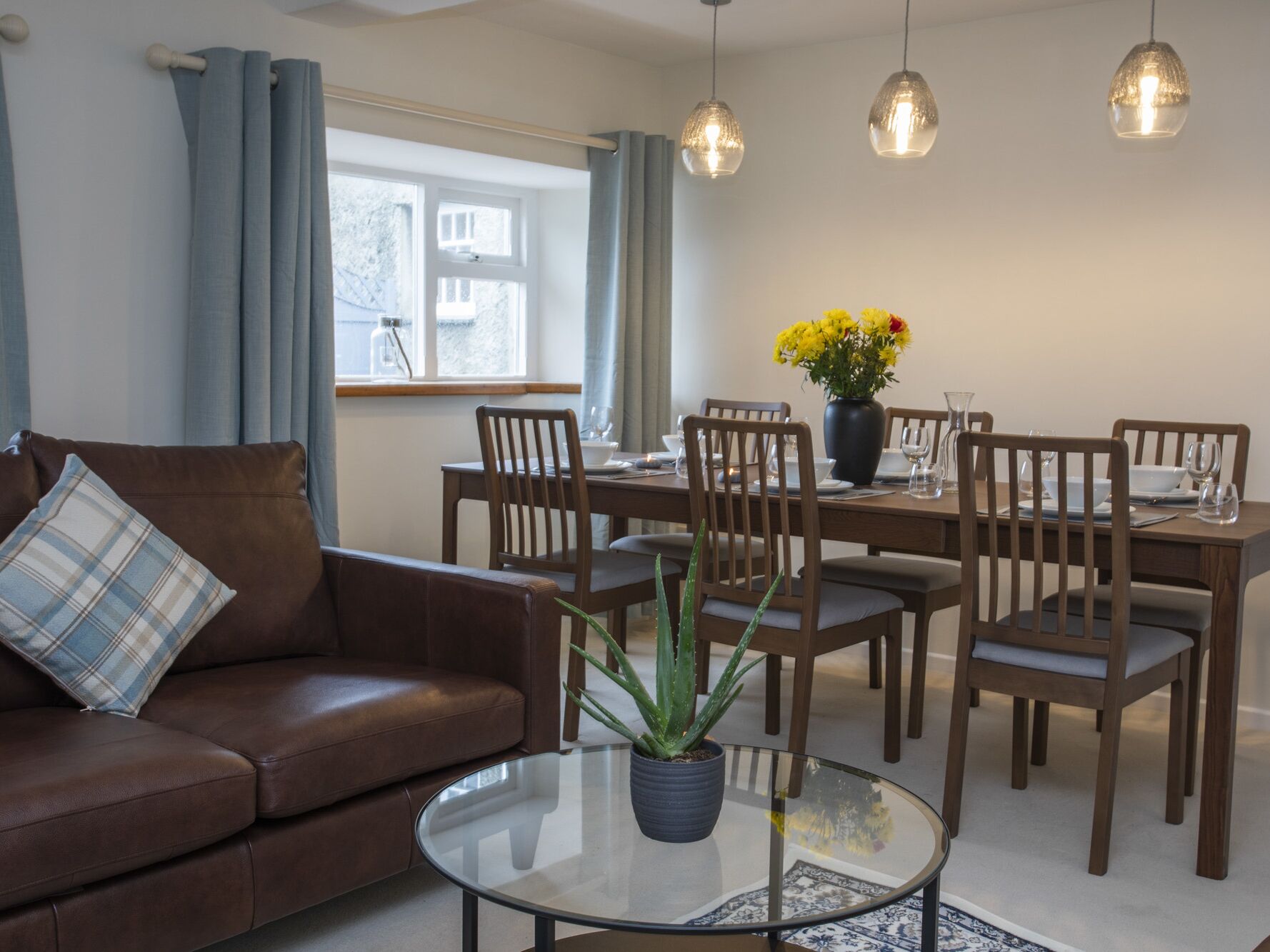 En moderne stue med en brun lædersofa, et sofabord i glas med en potteplante, træspisestole, et spisebord dækket til fire personer og hængende lamper.