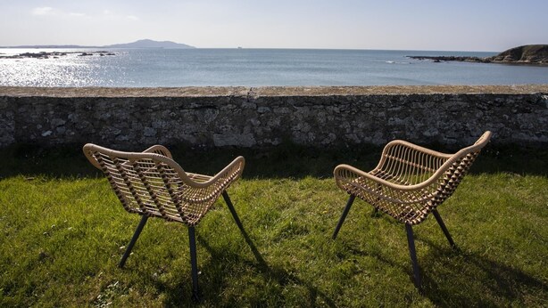 Two wicker chairs facing a stone wall overlooking a coastal view with a clear sky. - Stabl Bach (Holyhead)