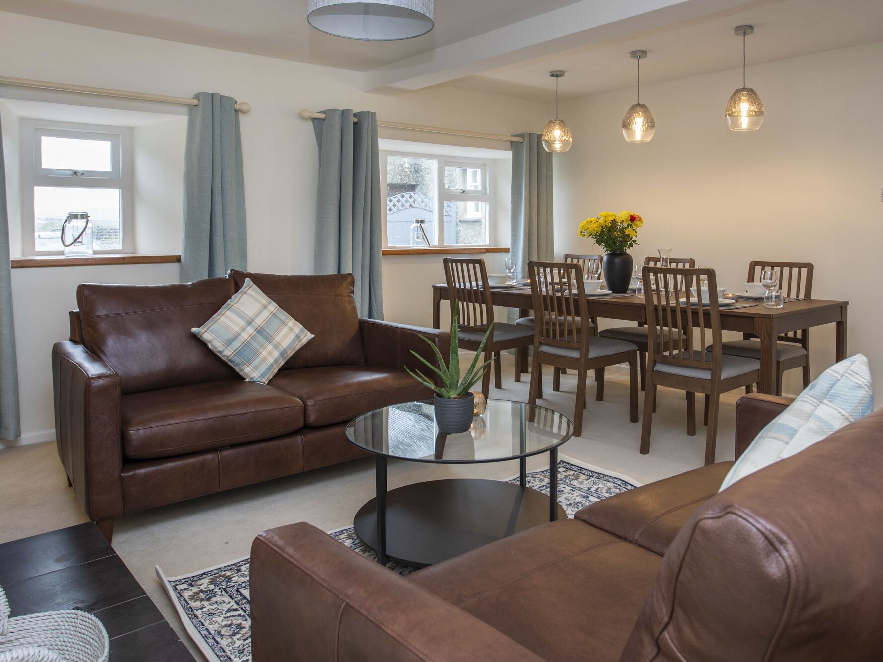 Un salon moderne avec un canapé en cuir marron, une table basse en verre et un coin repas comprenant des chaises et une table en bois, le tout disposé pour accueillir quatre personnes.