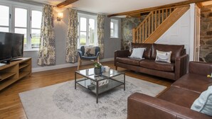 A cozy living room with a stone staircase, wooden beams, a television, a blue armchair, and a brown leather sofa. - Stabl Bach (Holyhead)
