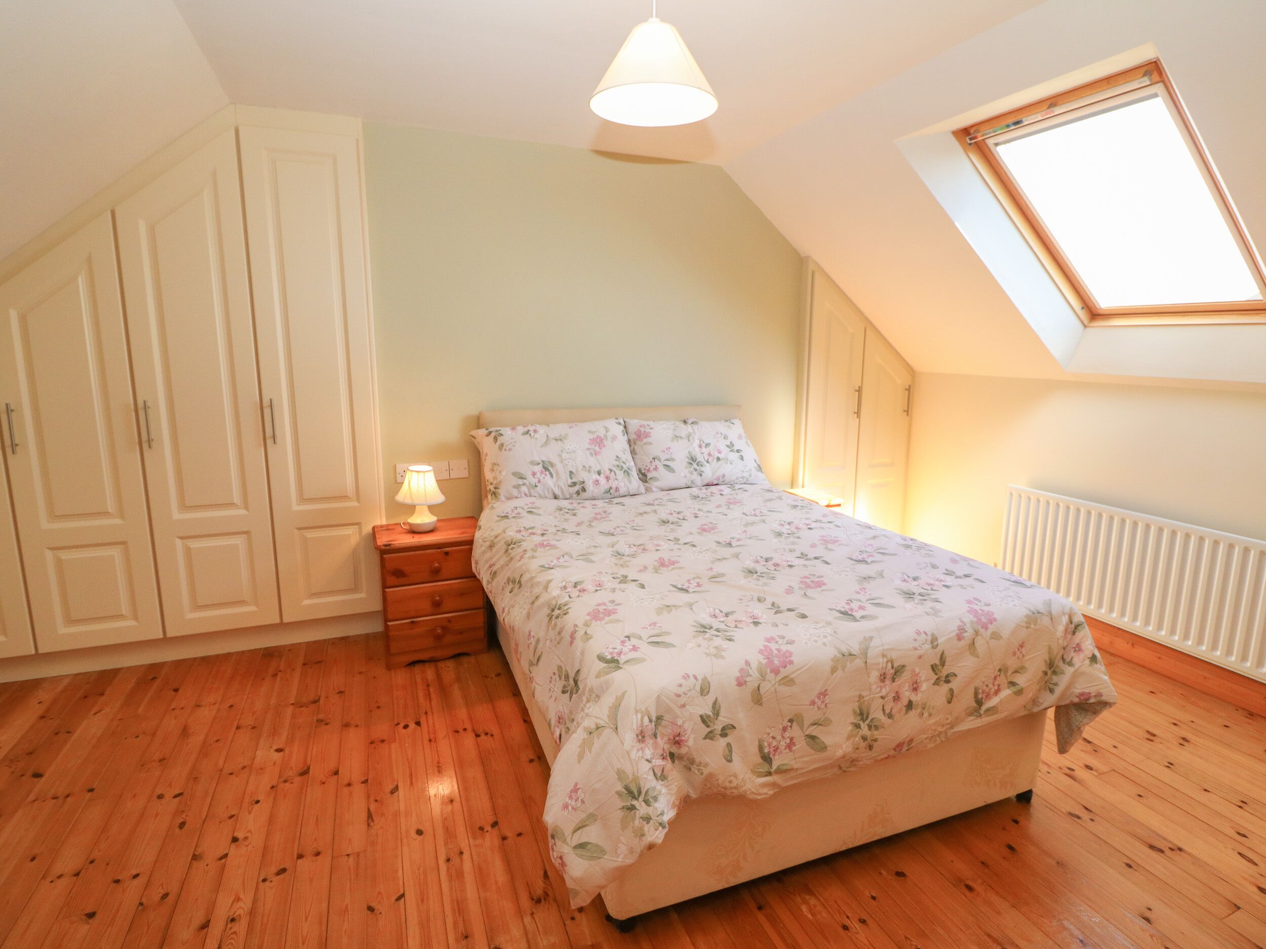 A bedroom with a wooden floor, a bed with floral bedding, a nightstand with a lamp, a wardrobe, and a skylight.