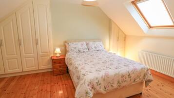 A bedroom with a wooden floor, a bed with floral bedding, a nightstand with a lamp, a wardrobe, and a skylight.