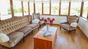 Una habitación con techo de madera y ventanas amplias, un sofá color beige, dos sillones y una mesa de centro de madera con revistas.