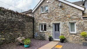A stone house with a grey door, a window with a sign reading "The Sanctuary", and a small patio area with potted plants and a pathway. - The Sanctuary (Windermere)