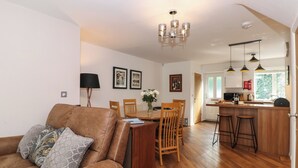A modern living room with a brown leather sofa, a dining table with chairs, a kitchen area with wooden cabinets and a bar, and a chandelier. - The Sanctuary (Windermere)