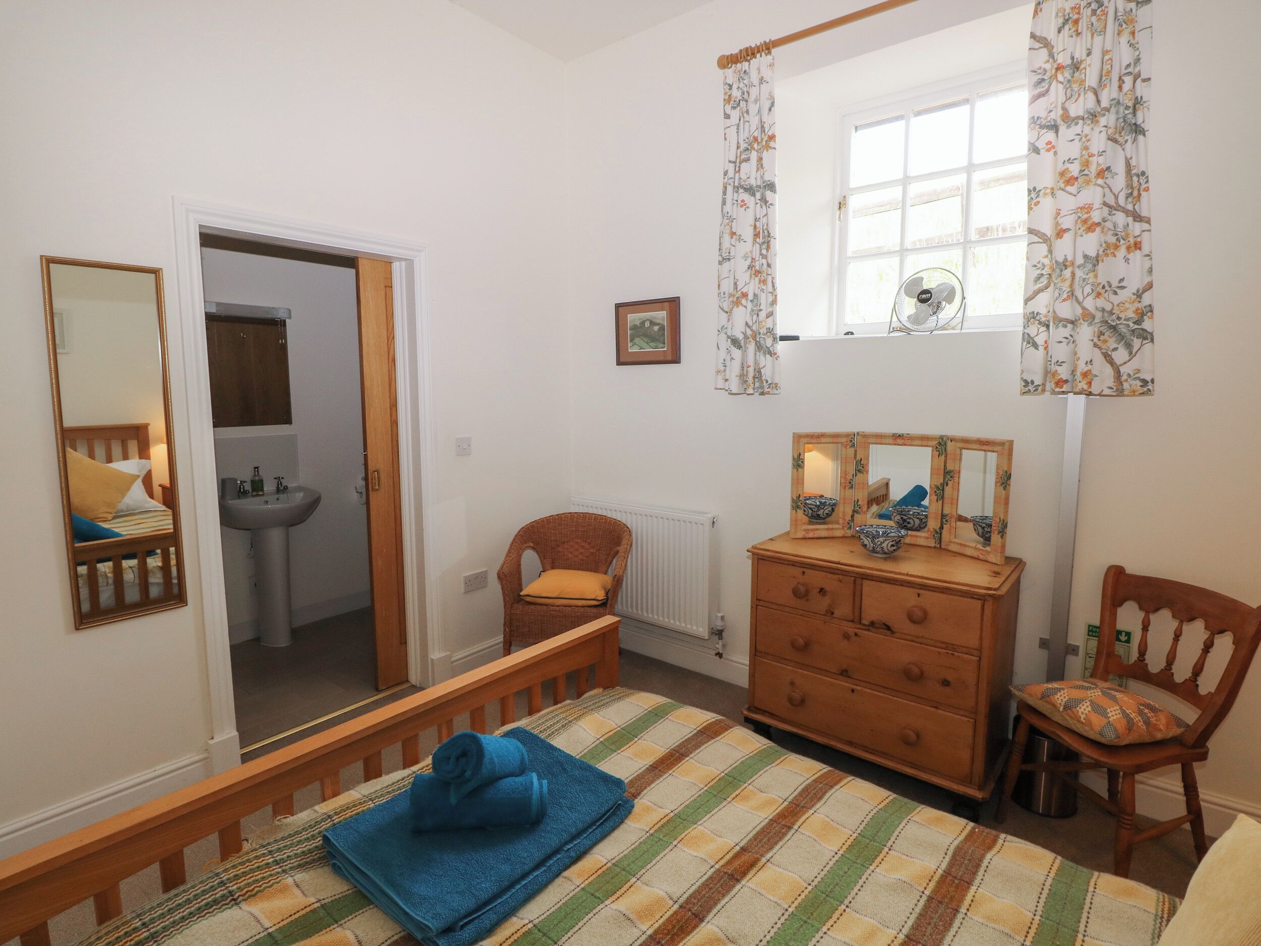 Quarto com cama, cômoda, cadeira, janela com cortinas, espelho e porta que dá para um banheiro.