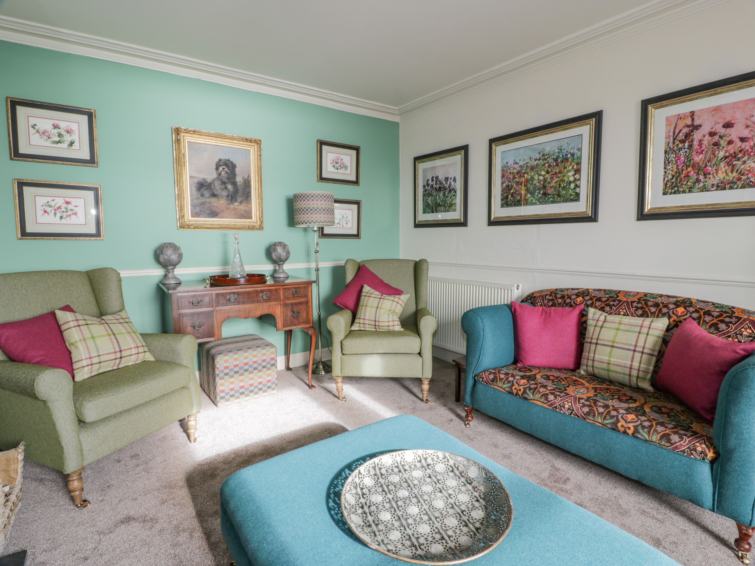 A living room with teal walls, green and blue sofas, and a patterned ottoman. There are framed pictures on the walls and a floor lamp.