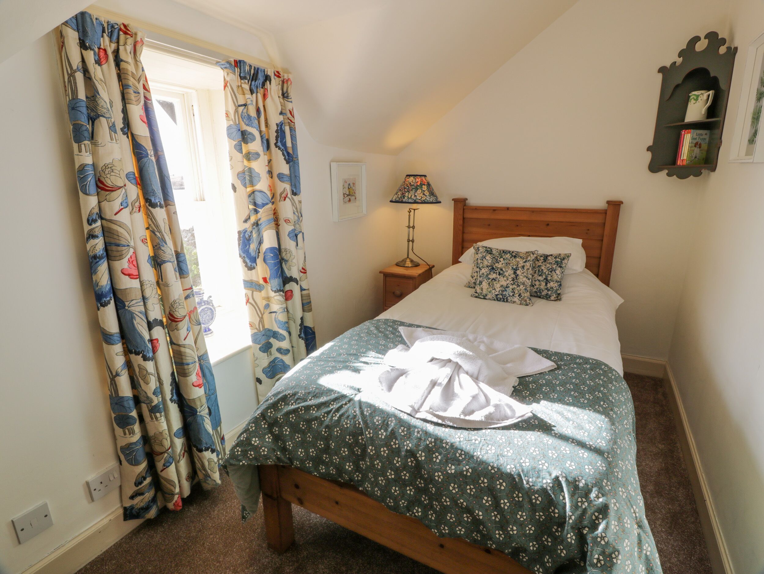 A bedroom with a bed, a nightstand, a lamp, a wall-mounted shelf, and a window with curtains.