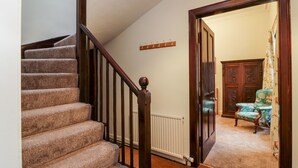 Una escalera con peldaños alfombrados y barandilla de madera, que conduce a una habitación con un radiador, una puerta de madera y un sillón con estampado floral.