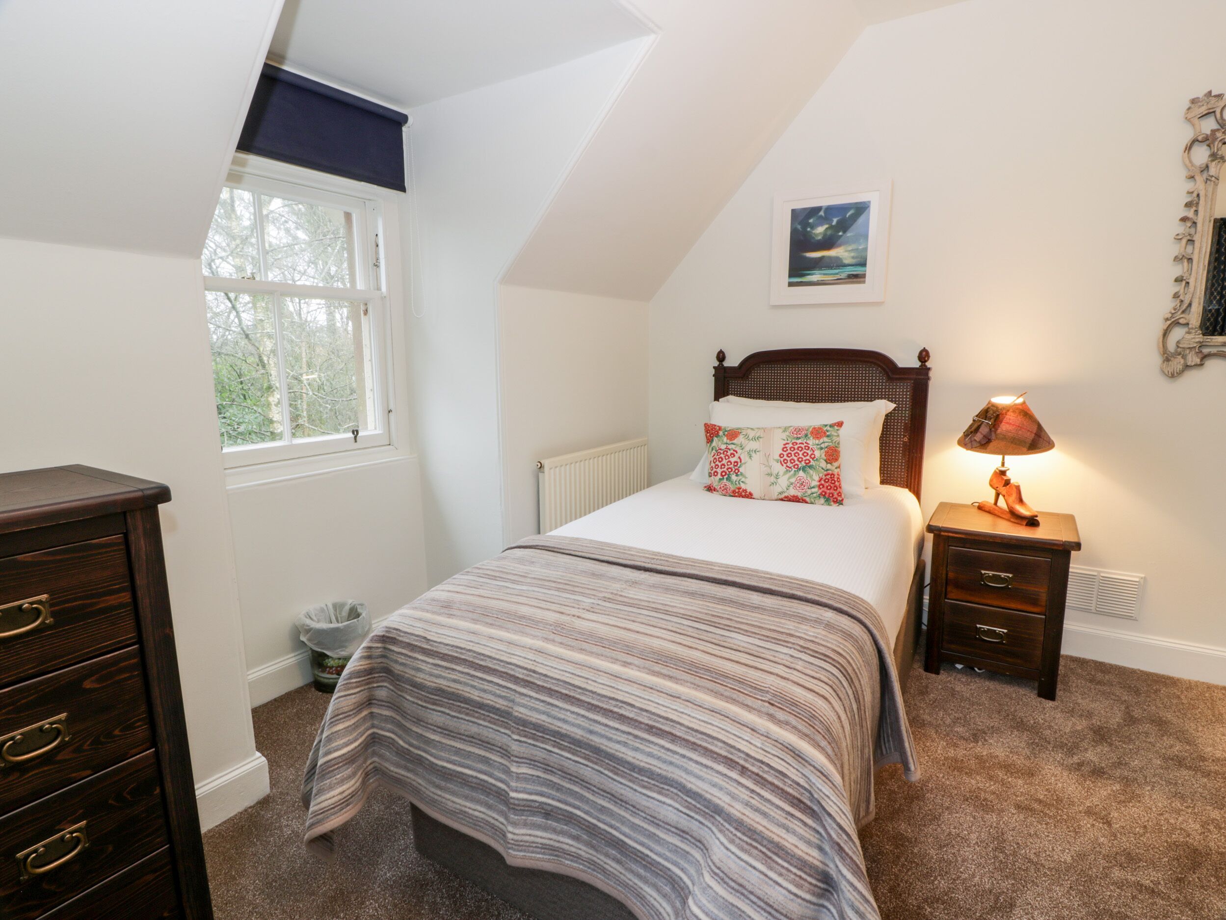 A bedroom with a bed, a dresser, a nightstand with a lamp, a window, and a picture on the wall.