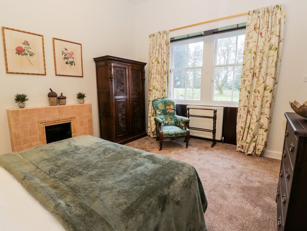 A bedroom with a bed, a chair, a fireplace, a large window with curtains, and two framed floral paintings on the wall. - Gate Lodge - Threave Estate (Castle Douglas)
