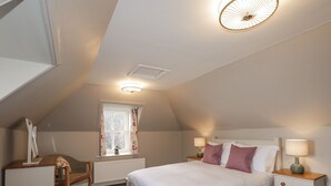 A bedroom with a sloped ceiling, a bed with white bedding, two bedside tables with lamps, a chair, and a window with curtains. - Craggan Cottage - Balmacara (Kyle)