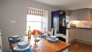 A dining area with a wooden table set for four, white chairs, a black refrigerator, and a window with a striped valance. - Craggan Cottage - Balmacara (Kyle)