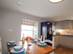 A dining area with a wooden table set for four, white chairs, a black refrigerator, and a window with a striped valance.
