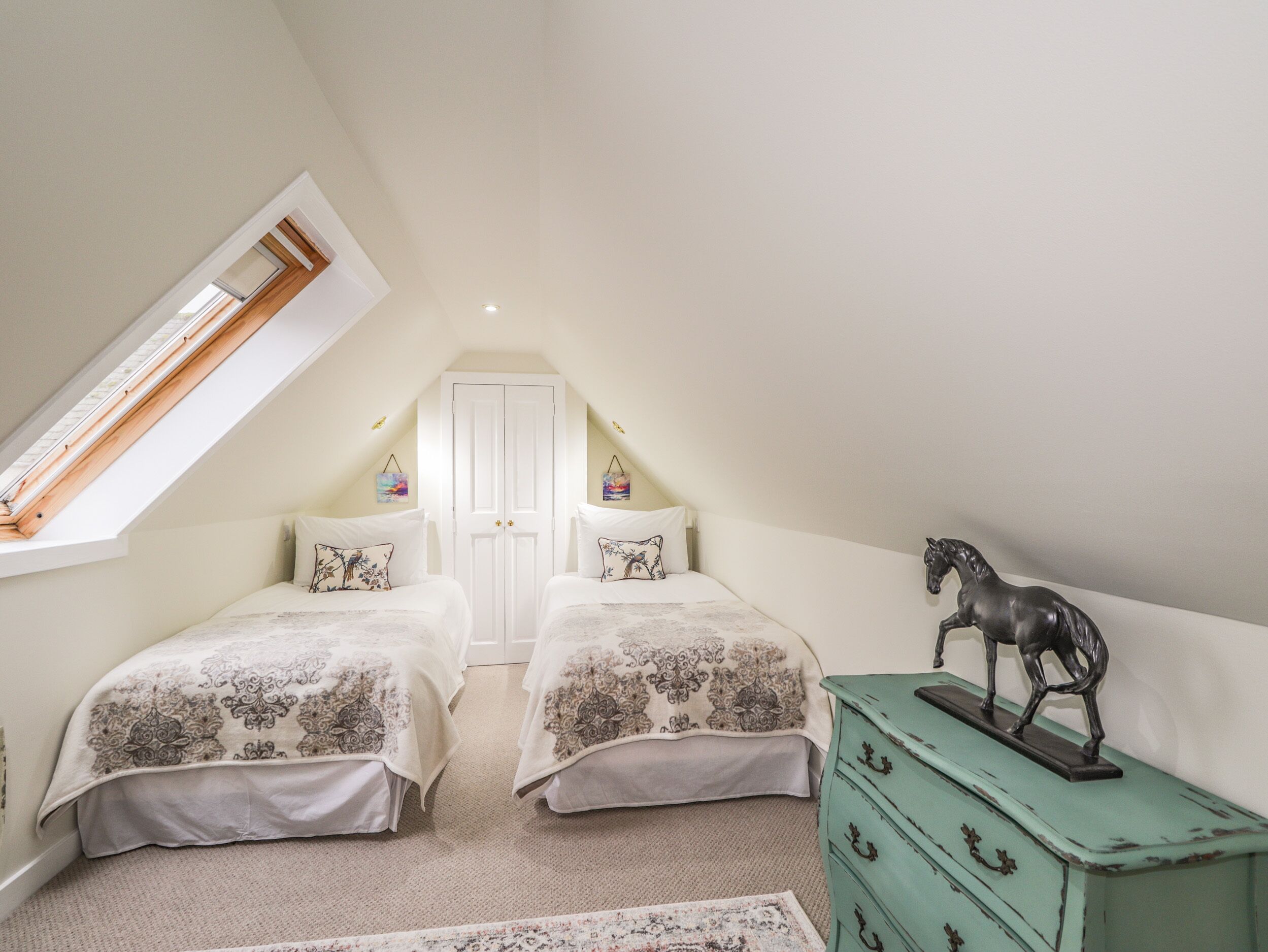 Une chambre avec deux lits, un puits de lumière, une porte blanche et une commode verte ornée d’une figurine de cheval.