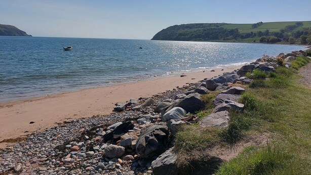 Una spiaggia con un litorale roccioso, acque calme e una collina in lontananza.
