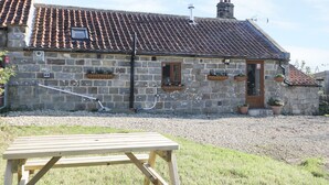 A stone building with a tiled roof, a wooden door, and a small window. There are flower boxes on the wall and a gravel driveway. - Dovecote Cottage (Scarborough)