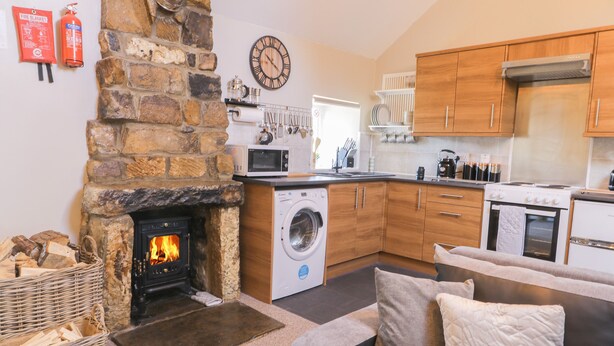 A cozy living room with a stone fireplace, a washing machine, and a kitchen with wooden cabinets and a stove. - Dovecote Cottage (Scarborough)