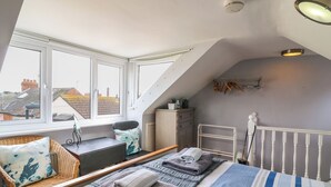 A bedroom with a sloped ceiling, large windows, a bed with a striped duvet, a bedside table, a chair, and a radiator. - Harbour Reach (Weymouth)