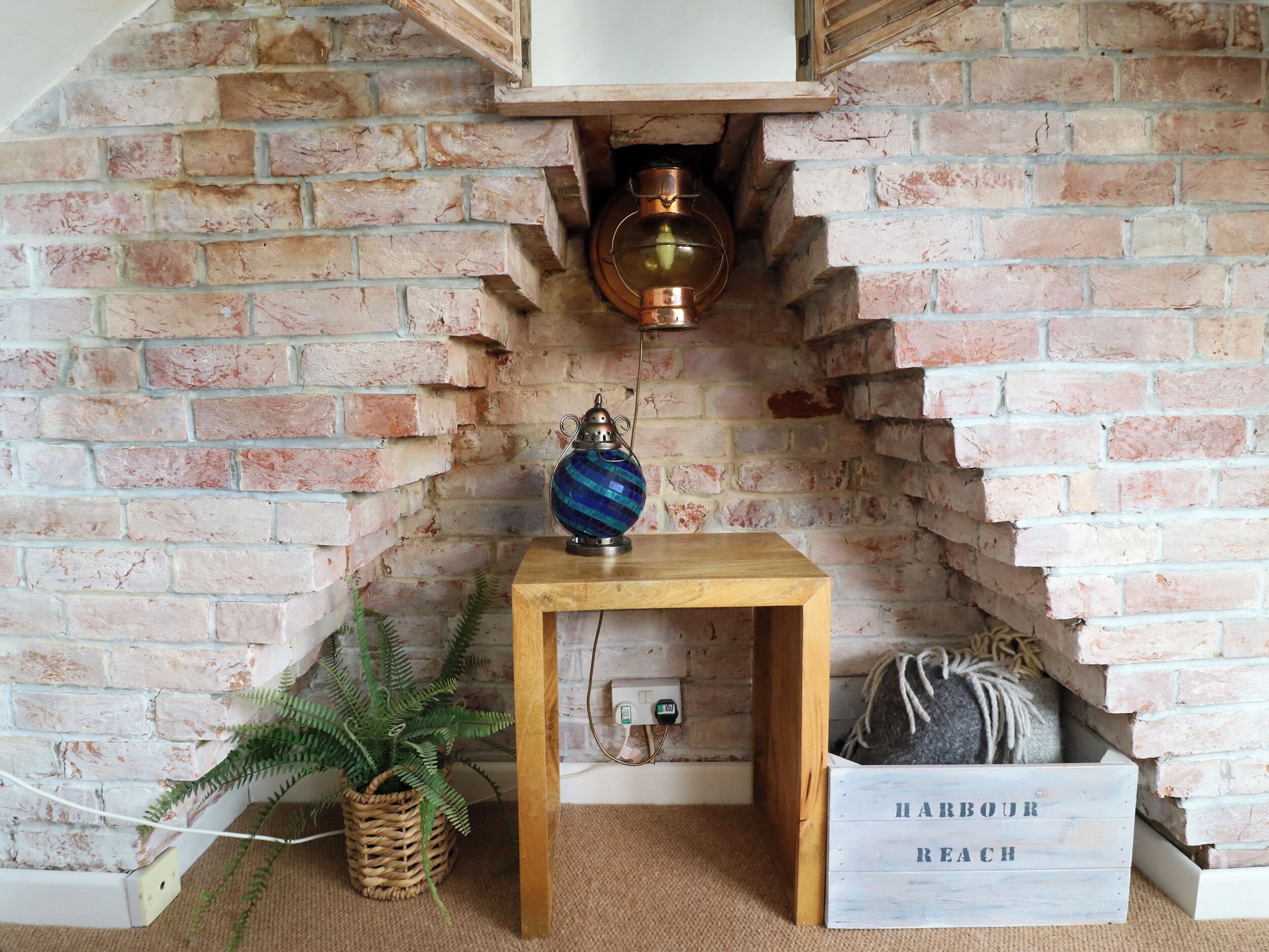 A rustic room with exposed brick walls, a wooden table with a globe lamp, a potted fern, and a wooden crate labeled "HARBOUR REACH".