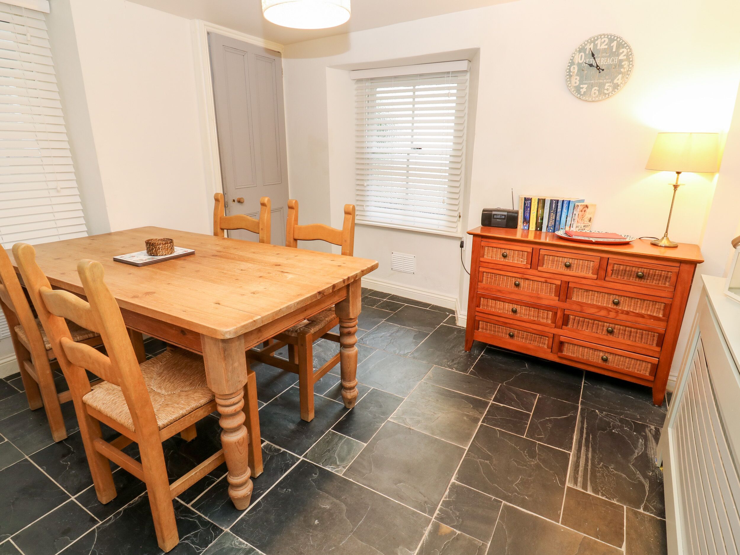Un coin repas avec une table et des chaises en bois, un buffet en osier contenant des livres et une horloge murale.