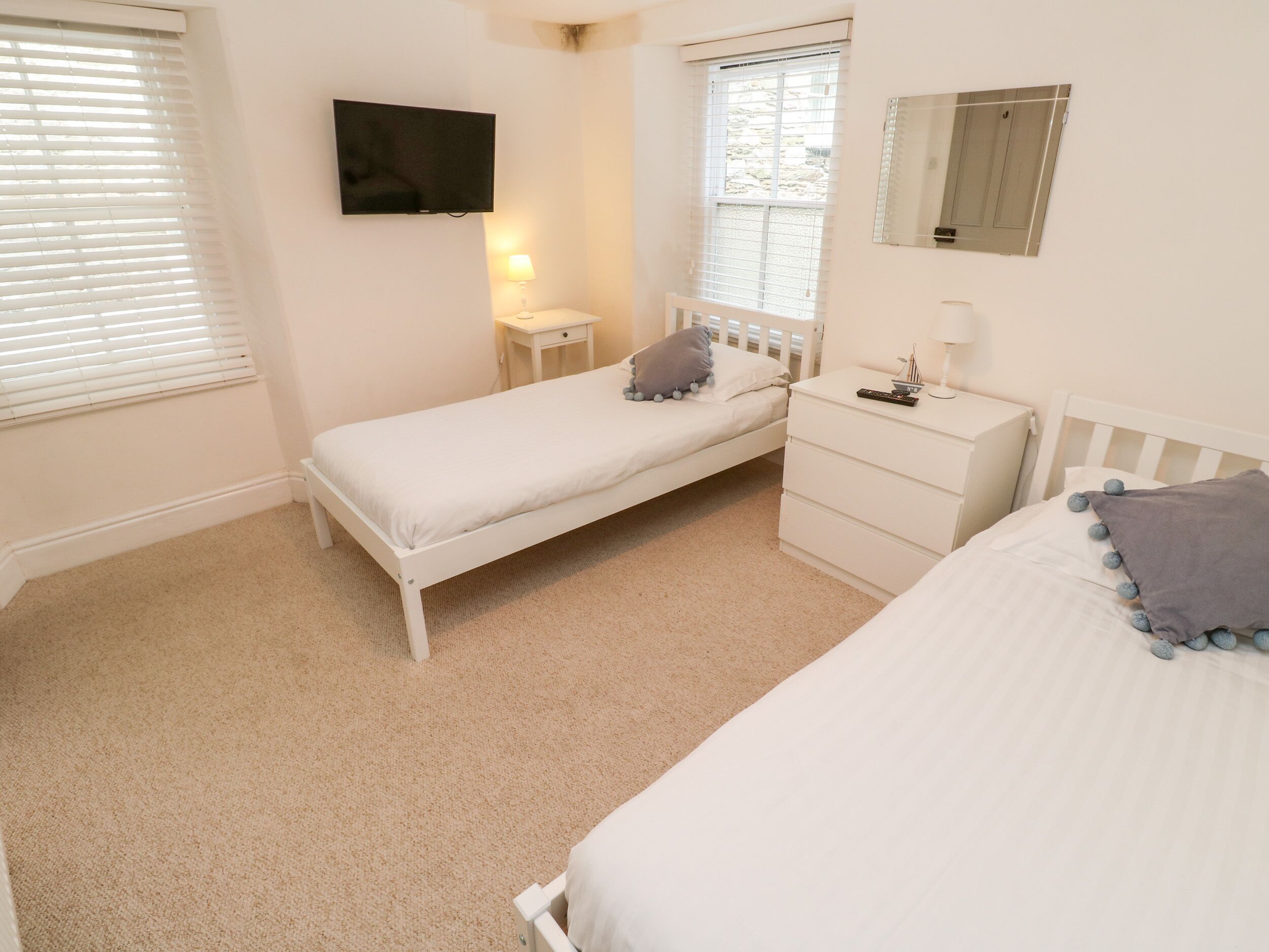 Une chambre avec un lit simple, une table de chevet avec une lampe, un téléviseur fixé au mur et une fenêtre avec des stores.